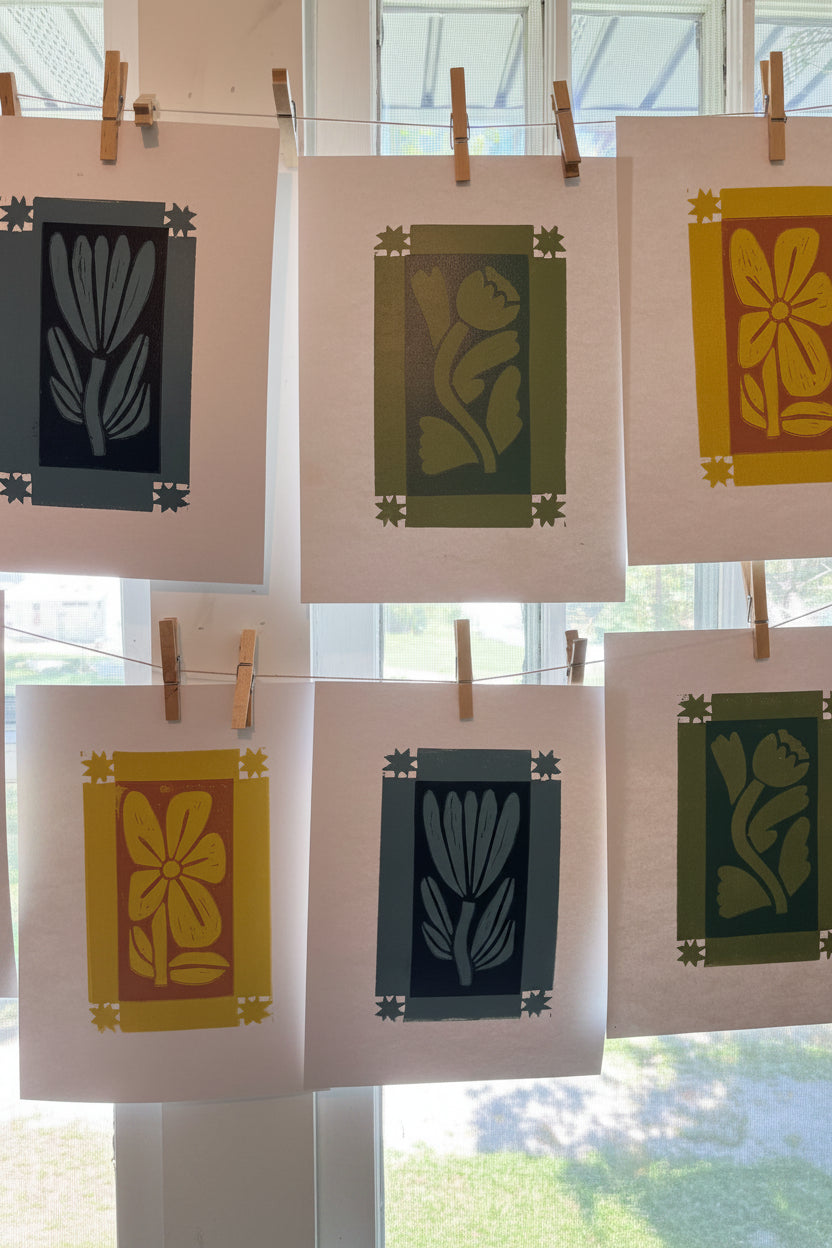 Six square art prints with floral and leaf motifs in yellow, green, and blue-gray tones, clipped to strings in front of a window.
